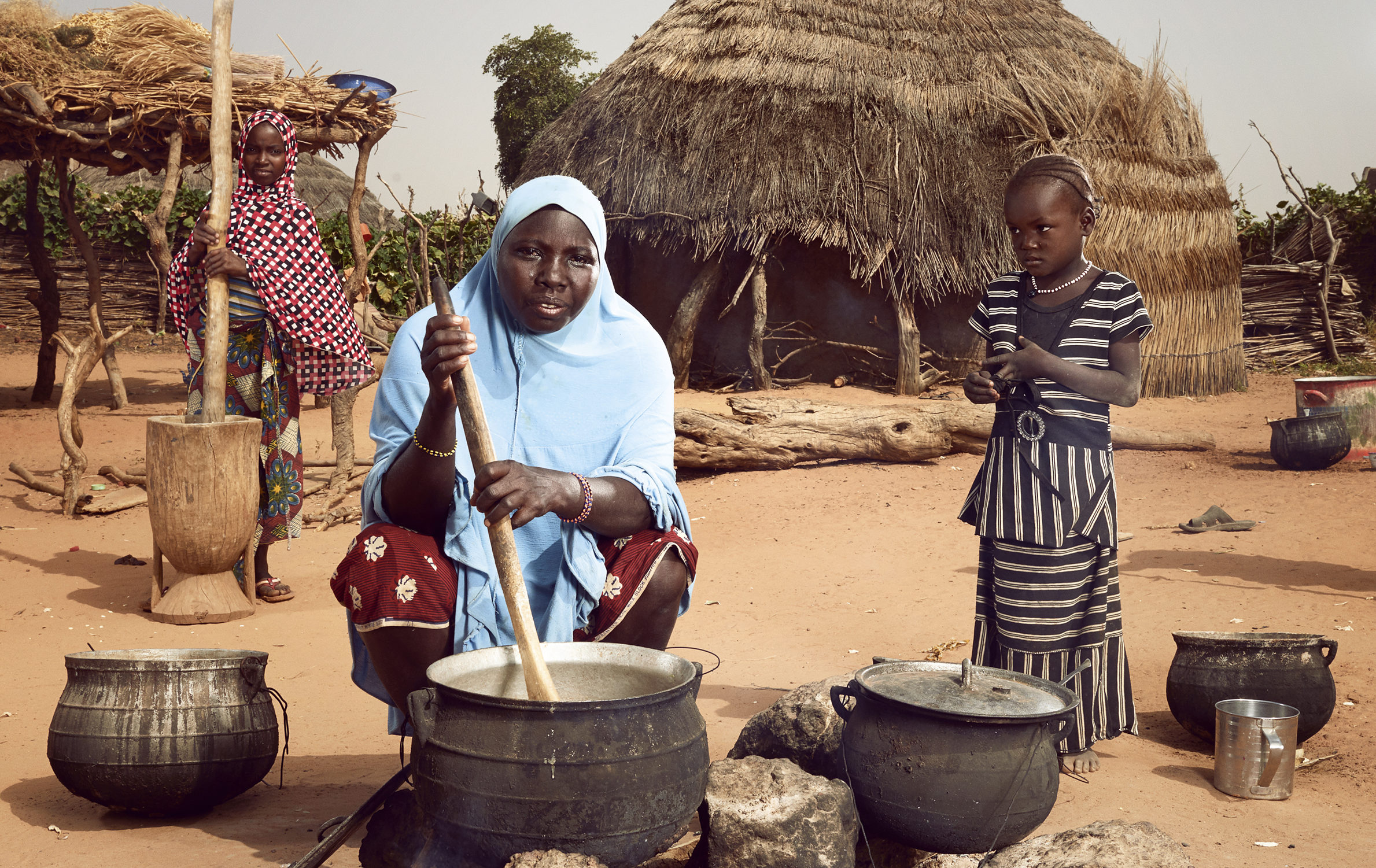 Hadiza (au centre) prépare un repas familial, aidée par ses filles Nadia, 4 ans, et Layhanatou, 10 ans, qui pilent le millet dans un mortier en bois. Photo : © Stephan Gladieu/Banque mondiale
